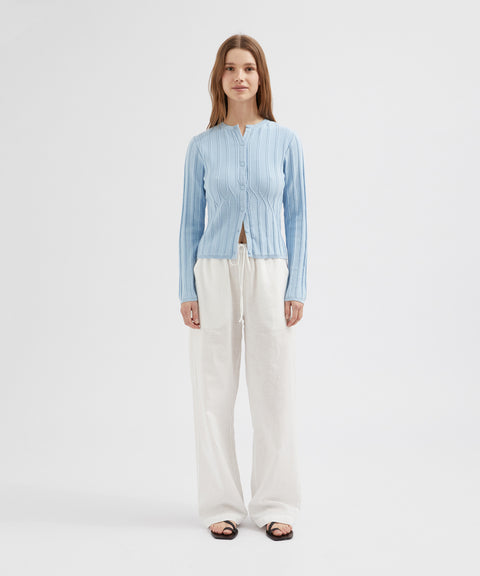 Woman wearing a light blue ribbed cardigan over a white top with white pants on a light gray background